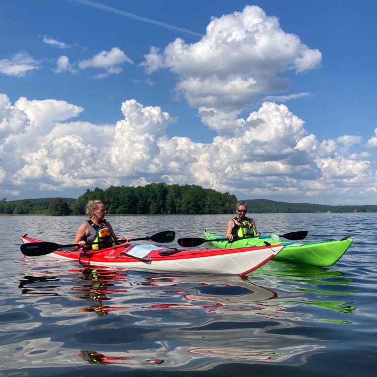 Två personer paddlar kajak på en sjö under en klar himmel med vita moln.