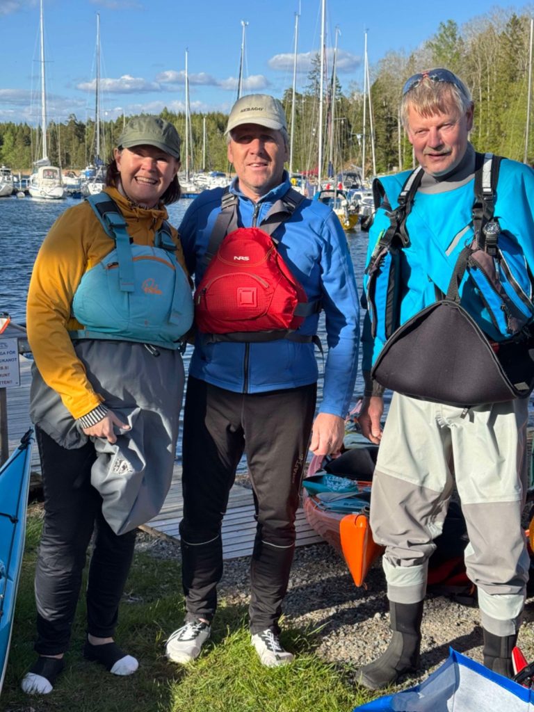 Tre personer i kajakutrustning vid en hamn, snart ska de paddla kajak. Soligt väder.