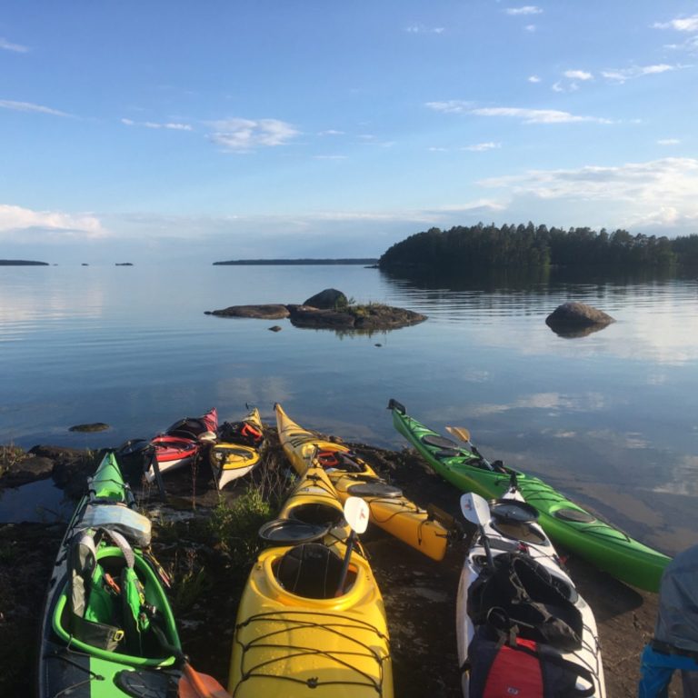 Flera kajaker i olika färger på stranden med stilla vatten och klippor i bakgrunden.
