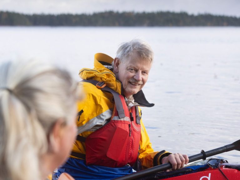 Två paddlare i kanot vid en sjö, en med grått hår och orange flytväst.
