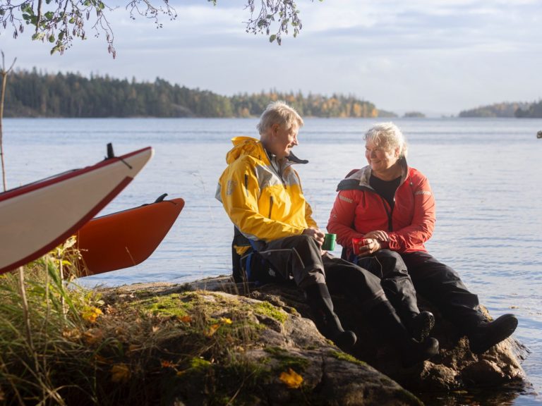 Två personer sitter vid vattnet, omgivna av natur, klädda i färgglada jackor och fikar.